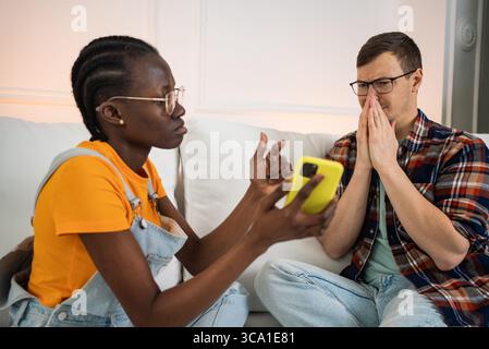 Junge Frau, die schockierende Nachrichten oder Fotos aus sozialen Medien auf dem Smartphone einem gestressten Mann zeigt, der zu Hause auf dem Sofa sitzt, bedeckt seinen Mund mit den Händen aus Angst oder Angst Stockfoto