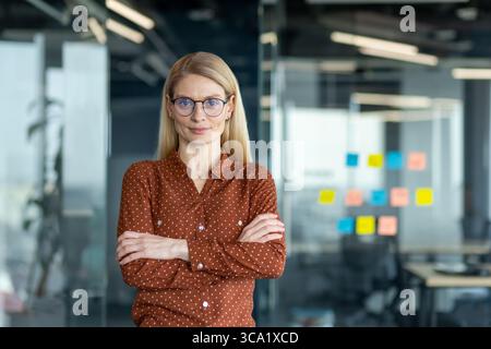 Eine blonde Frau in Brille mit überkreuzten Armen steht in einem Büro mit selbstbewusstem Ausdruck. Stockfoto