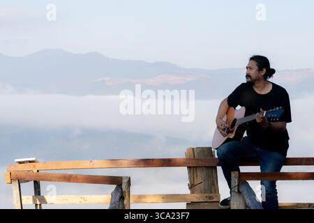 Junger Mann, der einen ruhigen Morgen genießt und Gitarre auf einem Holzbalkon mit warmem Sonnenlicht und malerischem Blick auf die Berge spielt. Stockfoto