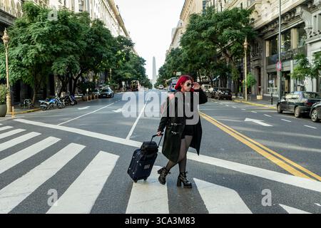 Junge Frau mit rotem Haar und Sonnenbrille, die den Koffer zieht und auf einer Fußgängerüberfahrt in buenos aires mit Obelisken im Hintergrund läuft Stockfoto