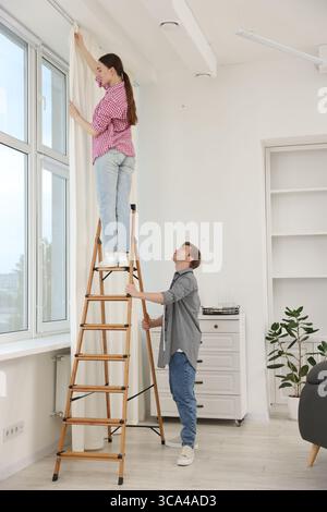 Ein Paar, das Vorhänge mit einer Leiter im Zimmer aufstellt Stockfoto