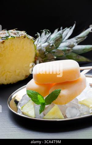 Ananassorbet-Eis-Eis-Eis auf Silberplatte mit Eiswürfeln gehackte Ananasscheiben, Minze nahe in halbe Ananas auf schwarzem Hintergrund geschnitten. Ananassorbet-Eis-Eis. Vertikal. Stockfoto