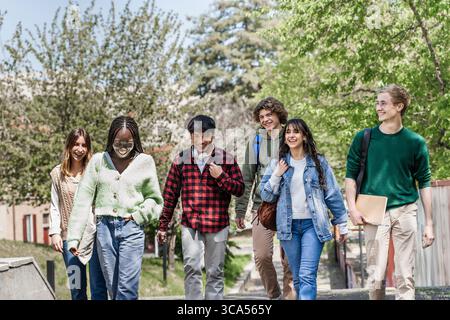 Energiegeladene und vielfältige Gruppe junger College-Freunde, die an einem sonnigen Tag gemeinsam auf einem Universitätscampus oder einem Parkweg im Freien lachen und spazieren. Jugend, Stockfoto