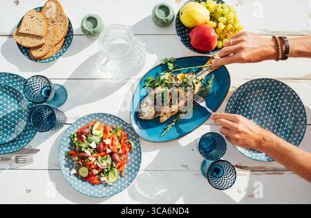 Goldbrasse (Orata) vom Grill gegrillt, serviert von Frau auf weißem Holztisch mit Gemüsesalat und Obst. Gesunde Meeresfrüchte-Mahlzeit, frisches Kochen Stockfoto