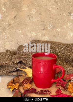 Eine warme Herbstszene mit einem roten Keramikbecher Kaffee oder Tee auf einer rustikalen Holzscheibe, umgeben von bunten Herbstblättern und einem gemütlichen Pullover Stockfoto