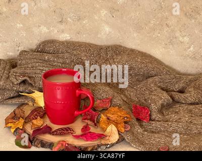 Eine warme Herbstszene mit einem roten Keramikbecher Kaffee oder Tee auf einer rustikalen Holzscheibe, umgeben von bunten Herbstblättern und einem gemütlichen Pullover Stockfoto