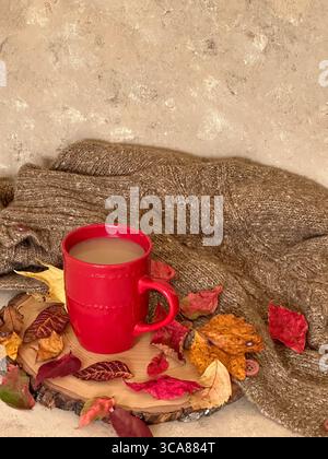 Eine leuchtend rote Kaffeetasse mit cremigem Kaffee liegt auf einer Holzplatte, umgeben von bunten Herbstblättern und einem warmen Strickpullover Stockfoto