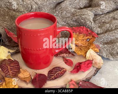 Eine warme Herbstszene mit einer roten Kaffeetasse gefüllt mit Kaffee, umgeben von bunten Herbstblättern, einer Strickdecke und rustikalem Holz. Stockfoto