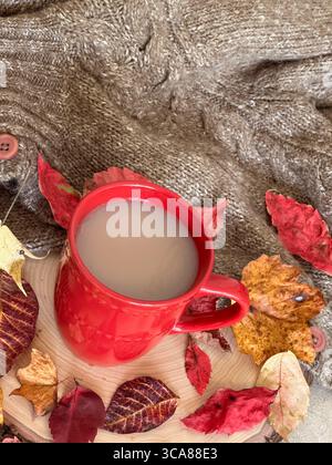 Eine warme Herbstszene mit einer roten Kaffeetasse gefüllt mit Kaffee, umgeben von bunten Herbstblättern, einer Strickdecke und rustikalem Holz. Stockfoto