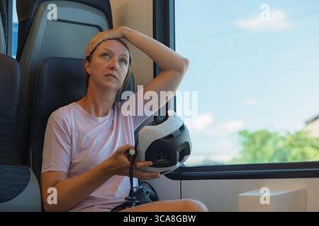 Eine Frau sitzt in einem Zug mit Fahrradhelm in den Händen und sieht nachdenklich aus, während sie aus dem Fenster auf einen sonnigen Tag blickt. Stockfoto
