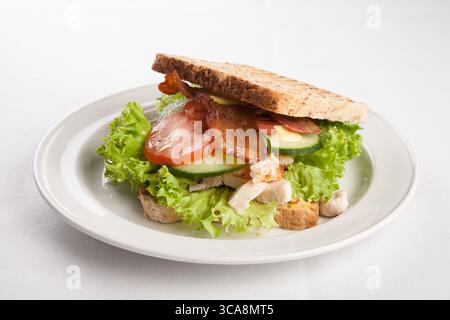 Getoastetes Sandwich auf einem weißen Teller gefüllt mit Salat, Speck, Gurke und Huhn, eine klassische und appetitliche Mahlzeit, Studio-Food-Shot Stockfoto