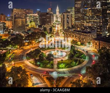 Ein lebhaftes nächtliches Stadtbild zeigt ein gut beleuchtetes Stadtgebiet mit einem prominenten runden plaza und einem Brunnen, bekannt als Logan Square in Philadelphia, Pen Stockfoto