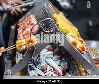 Gegrillte thailändische Würstchen auf Spießen über heißer Holzkohle in der Nacht, die das Wesen der lebendigen Street Food-Kultur einfangen. Stockfoto