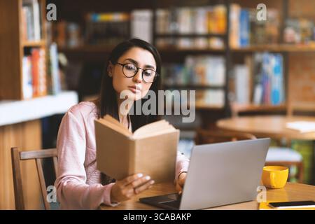 Nachdenkliche lateinerin liest vor dem Laptop in der Cafeteria ein Buch, studiert und bereitet sich auf die Prüfung vor. Stockfoto