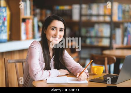Begeisterter lateinamerikanischer Student, der online einen Job sucht, mit Laptop und Notizen macht, im Café sitzt, Kopierraum Stockfoto
