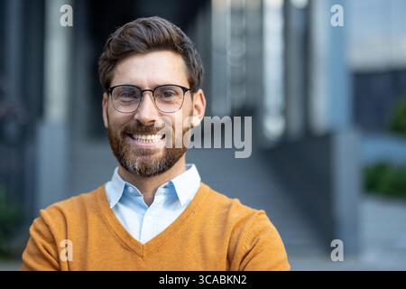 Bärtiger Mann mit Brille in lässigem Outfit, der Selbstvertrauen und Positivität in einer modernen urbanen Umgebung mit verschwommenem Hintergrund ausstrahlt. Stockfoto