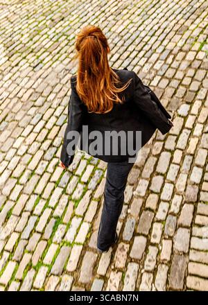 Eine Person mit langen roten Haaren läuft auf einer kopfsteingepflasterten Straße. Stockfoto