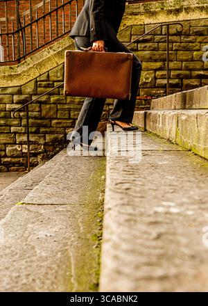 Eine Person in Anzug und Fersen geht eine Steintreppe hinauf, während sie eine Aktentasche trägt. Stockfoto