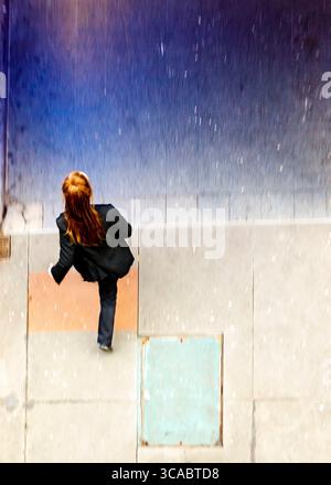 Eine Frau mit langen roten Haaren, die einen schwarzen Anzug trägt, läuft im Regen einen Bürgersteig hinunter. Stockfoto