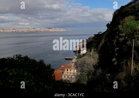 Sonnendurchflutete Klippen und historische Häuser von Praia das Lavadeiras in Almada überblicken Lissabons Fluss Tejo und verschmelzen Küstenschönheit mit lebhaftem urbanem Charme. Stockfoto