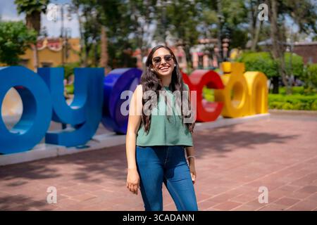 Die glückliche junge Latina posiert selbstbewusst vor dem farbenfrohen Coyoacan-Schild in Mexiko-Stadt, trägt eine Sonnenbrille, ein lässiges ärmelloses grünes Oberteil und eine jeans Stockfoto