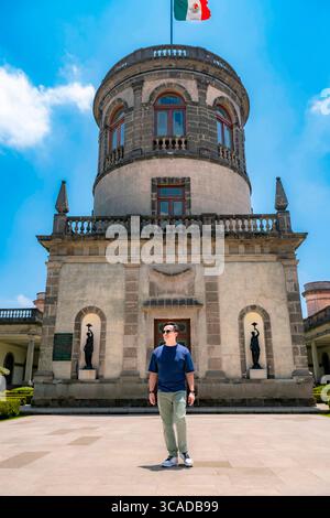 Glücklicher junger Latino-Mann mit marineblauem Hemd, hellgrüner Hose und Sonnenbrille posiert stolz vor dem historischen Turm von Chapultepec Castle, Mexiko Stockfoto