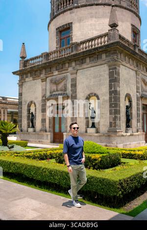 Glücklicher junger Latino-Mann mit marineblauem Hemd, hellgrüner Hose und Sonnenbrille posiert stolz vor dem historischen Turm von Chapultepec Castle, Mexiko Stockfoto