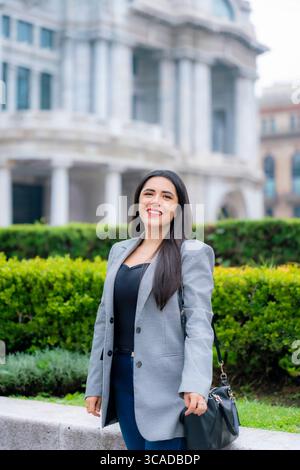 Junge lächelnde Geschäftsfrau Latina mit langen glatten Haaren, grauem kariertem Blazer, dunklem Oberteil und blauer Hose, die fröhlich im Palacio de Bellas AR posiert Stockfoto