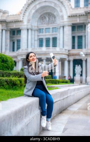 Selbstbewusste, glückliche junge Latina Geschäftsfrau, die lässig sitzt und lächelt, während sie ein Selfie in grauem Blazer und blauer Hose im Palacio de Bellas Artes macht. Stockfoto