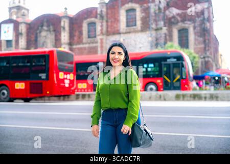Lächelnde junge, glückliche Latina-Frau posiert in der Innenstadt von Mexiko-Stadt vor dem roten Metrobus-System, trägt grüne Bluse und Jeans mit dem Heritage-Bogen Stockfoto