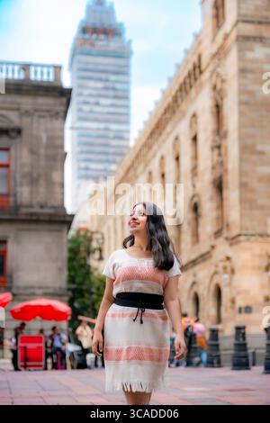 Fröhliche junge Latina in einem weißen, korallengewobenen Kleid mit schwarzem Schärpe spaziert lächelnd durch die Innenstadt von Mexiko-Stadt in der Nähe des Torre Latinoamericana Stockfoto