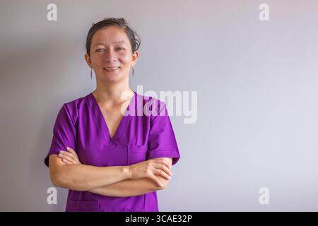Porträt einer lächelnden mittelerwachsenen Krankenschwester, die lila Peelings trägt, mit überkreuzten Armen vor einem schlichten Hintergrund steht, Selbstvertrauen und Geständnis ausstrahlt Stockfoto
