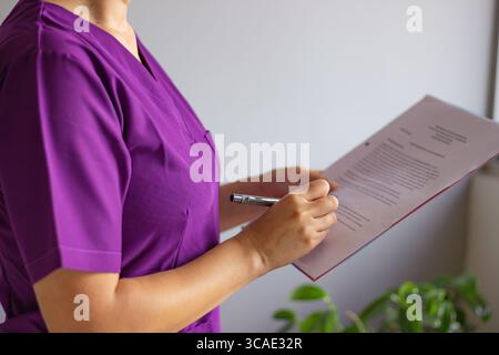 Weibliche Krankenschwester in lila Uniform liest und unterzeichnet ein medizinisches Dokument oder eine Akte, hält einen Stift in der Hand, steht in einer medizinischen Praxis Stockfoto