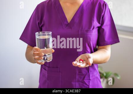 Weibliche Ärztin, die lila Peelings trägt, hält ein Glas Wasser und Medikamente, bereit, Pillen an einen Patienten zu verabreichen, der behandelt werden muss Stockfoto