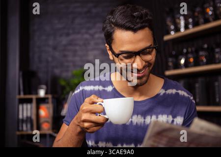 Mann mit weißem Becher und Zeitung in gemütlichem Café mit Backsteinmauern Hintergrund Stockfoto