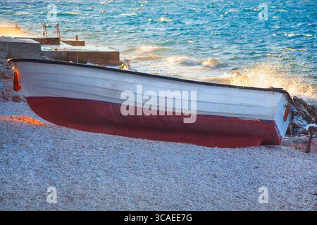 Verwittertes weißes Boot ruht am Kiesstrand. Wellen krachen gegen die Küste und stoßen sanft ein kleines, weißes Boot auf den Strand Stockfoto