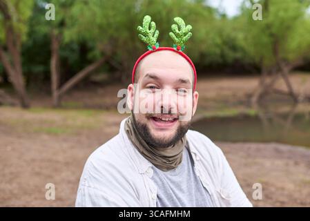 Ein junger hispanischer Mann, der ein weihnachtliches Rentiergeweih trägt, lächelt draußen in einer natürlichen Umgebung in die Kamera. Stockfoto