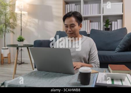 Ein junger Mann sitzt zu Hause auf dem Boden, arbeitet an seinem Laptop und denkt ernsthaft nach. Er konzentriert sich auf seine Online-Aufgaben, das Lernen oder die Arbeit aus der Ferne Stockfoto