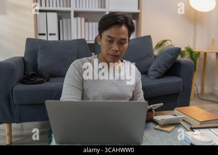 Ein junger Mann arbeitet von zu Hause aus, sitzt auf dem Boden mit seinem Laptop und Smartphone, umgeben von Notizbüchern, Dokumenten und Kaffee. Er scheint fokussiert zu sein Stockfoto