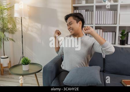 Ein junger Mann in einem grauen Hemd entspannt sich und dehnt seine Arme bequem, während er zu Hause auf einem gemütlichen blauen Sofa sitzt. Er genießt einen friedlichen Moment, lächelt Stockfoto