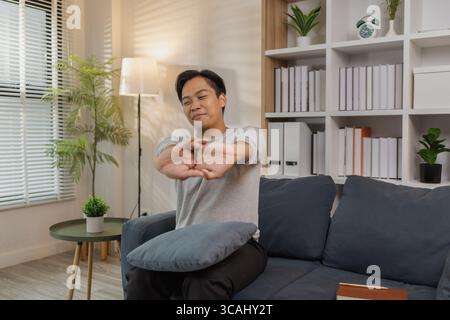 Ein junger Mann in einem grauen Hemd entspannt sich und dehnt seine Arme bequem, während er zu Hause auf einem gemütlichen blauen Sofa sitzt. Er genießt einen friedlichen Moment, lächelt Stockfoto
