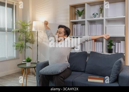 Ein junger Mann in einem grauen Hemd entspannt sich und dehnt seine Arme bequem, während er zu Hause auf einem gemütlichen blauen Sofa sitzt. Er genießt einen friedlichen Moment, lächelt Stockfoto