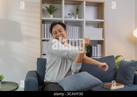 Ein junger Mann in einem grauen Hemd entspannt sich und dehnt seine Arme bequem, während er zu Hause auf einem gemütlichen blauen Sofa sitzt. Er genießt einen friedlichen Moment, lächelt Stockfoto