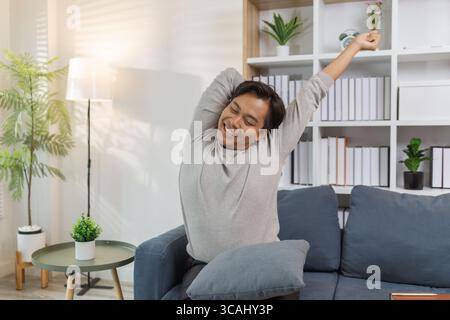 Ein junger Mann in einem grauen Hemd entspannt sich und dehnt seine Arme bequem, während er zu Hause auf einem gemütlichen blauen Sofa sitzt. Er genießt einen friedlichen Moment, lächelt Stockfoto