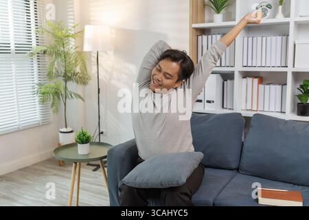 Ein junger Mann in einem grauen Hemd entspannt sich und dehnt seine Arme bequem, während er zu Hause auf einem gemütlichen blauen Sofa sitzt. Er genießt einen friedlichen Moment, lächelt Stockfoto