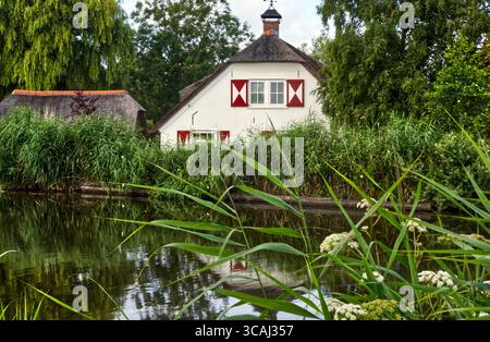 Traditionelle niederländische Polderwohnung mit Strohdach, roten Fensterläden, die sich friedlich im ruhigen Kanalwasser inmitten von grünem Land spiegeln Stockfoto