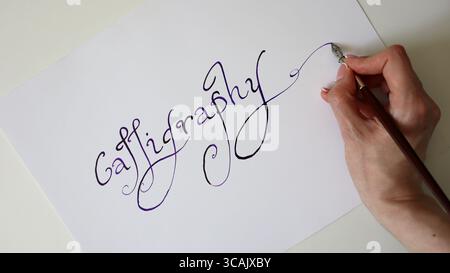 Der Kalligraphieprozess. Weibliche Hand schreibt das Wort Kalligraphie. Handgeschriebene Schriftart. Abgerundete Buchstaben, lange Reihen. Die Schriftart hat einen Farbverlauf von Schwarz zu Violett. Stockfoto