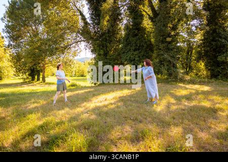 Kaukasische Mutter und Tochter werfen Frisbee und lächeln auf Gras im Stadtpark. Familienverbundenheit, echte Outdoor-Aktivitäten und gesunder Lebensstil im Urlaub Stockfoto