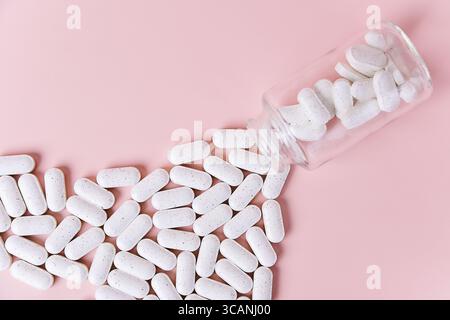 Eine Ansicht von oben nach unten zeigt weiße, längliche Pillen, die aus einer durchsichtigen Glasflasche auf einen rosafarbenen Hintergrund gelangen. Gesundheit, Wellness und Pharmazie Stockfoto