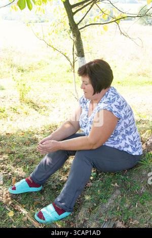 Eine ältere Frau mit dunklen Haaren sitzt im Kreuz unter einem jungen Baum auf einem grasbewachsenen Feld. Sie trägt ein Hemd mit Vogelmuster und eine graue Hose. Stockfoto